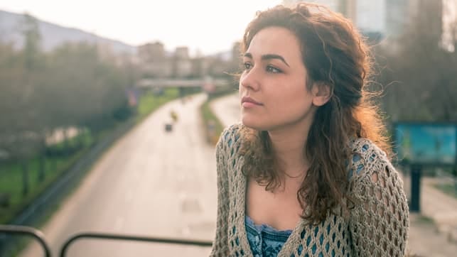 a young girl with dark curly hair is thinking while standing on a balcony in summer