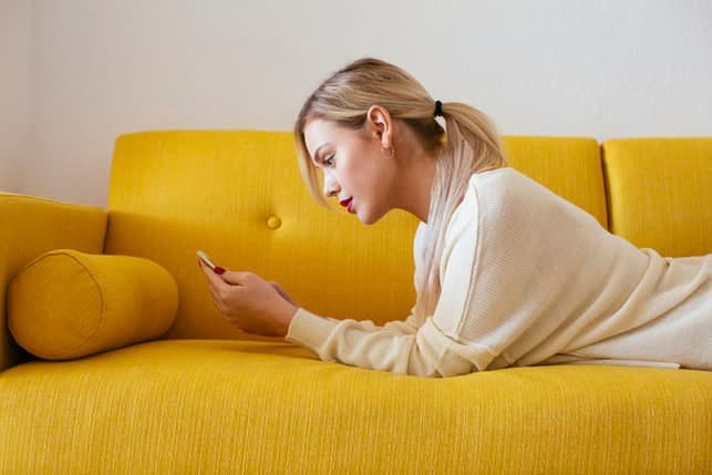 a blonde woman is checking her phone while lying on a yellow sofa