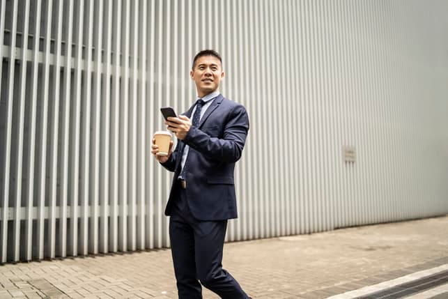 an asian businessman is checking his phone outdoors while drinking his coffee